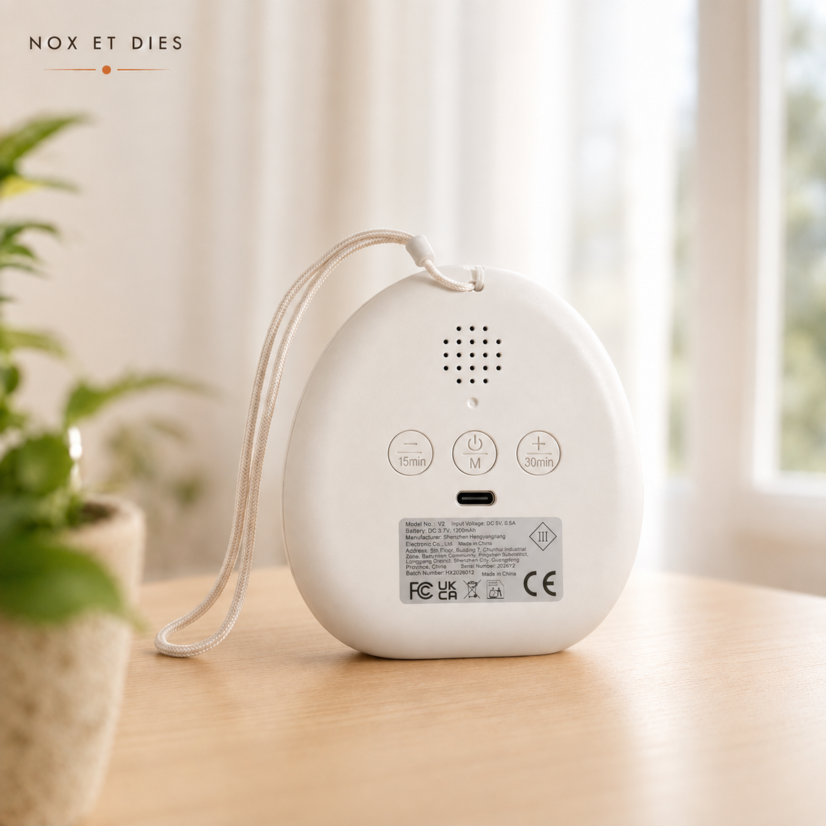 Vue arrière de la veilleuse portable Bruma Nox et Dies posée sur un meuble en bois, montrant les commandes tactiles, le port de charge Type-C et la lanière, avec une plante en fond flou