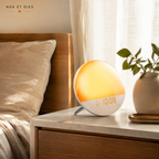 Réveil simulateur d'aube connecté Demi Wifi Nox et Dies posé sur une table de chevet, lumière chaude orangée allumée, écran affichant l'heure